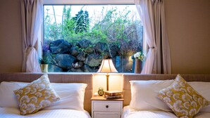 A double bed with matching pillows, a bedside table with a lamp, and a view of rocks and greenery through a window. - Villa Hanasaku Fujikawaguchiko B (Fujikawaguchiko)