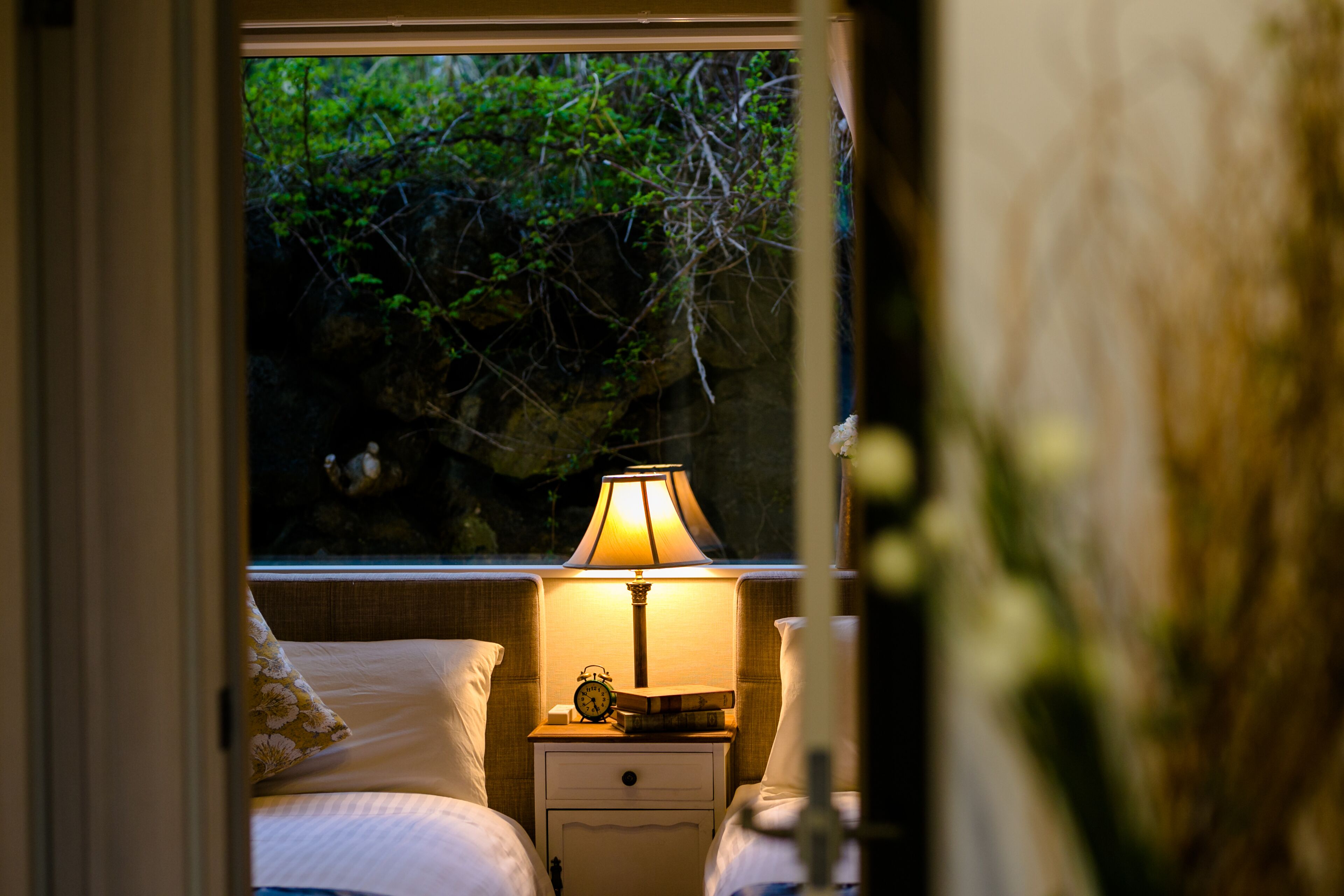 Ein Schlafzimmer mit Nachttisch, Nachttischlampe und einem Fenster mit Blick auf Grünflächen.