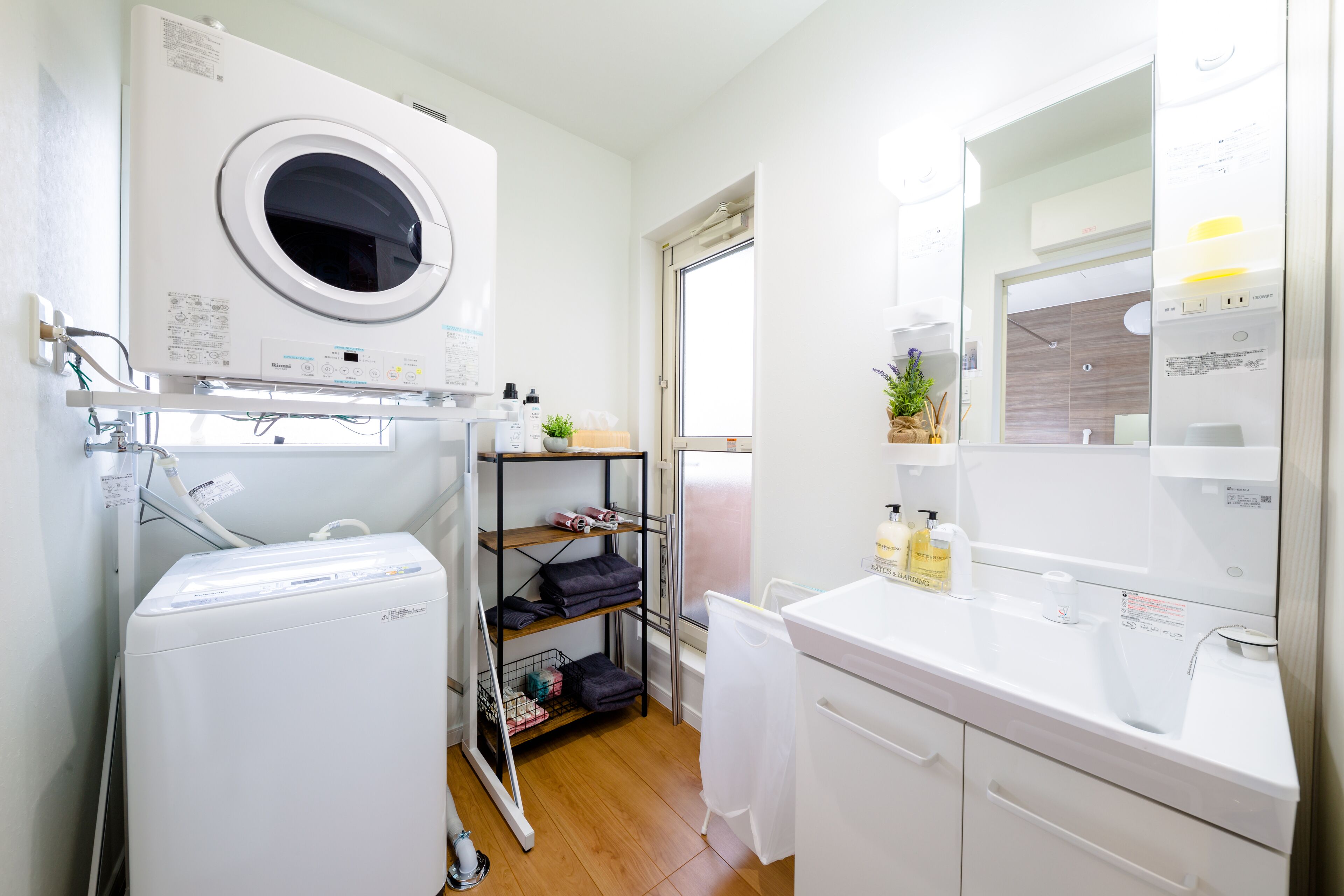 A compact hotel bathroom with a washing machine, a shelf with folded towels, a mirror, and a sink.