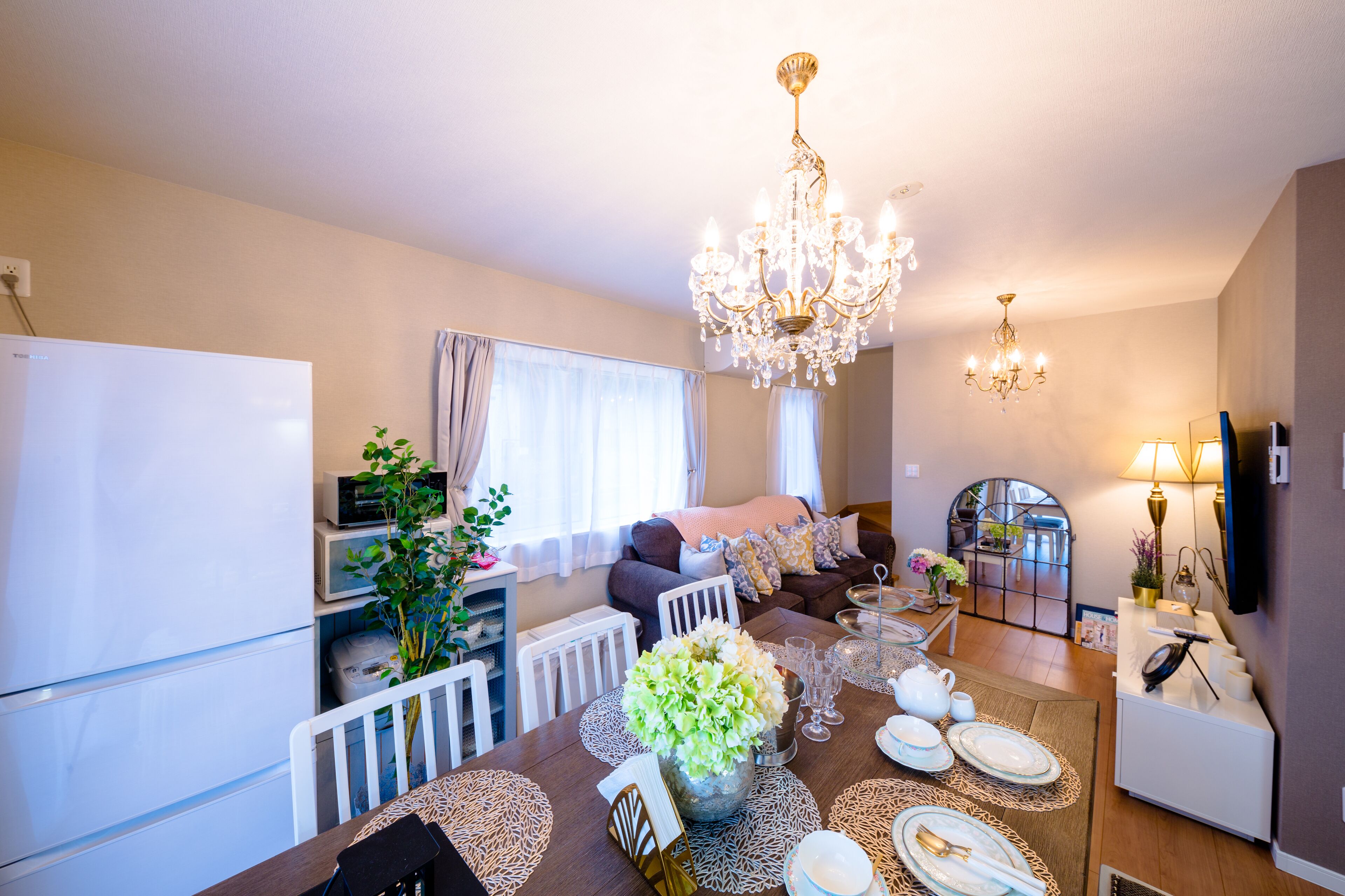 A modern kitchen with a dining area, a chandelier, and a living room with a sofa and a TV.