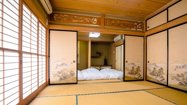 Une pièce de style japonais traditionnel, avec un sol recouvert de tatamis, des portes coulissantes décorées et un lit avec une literie blanche.