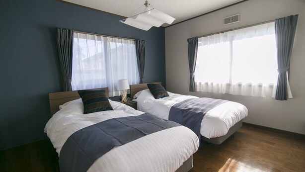 Ein Hotelzimmer mit zwei Betten, einem Nachttisch, einer Lampe und großen Fenstern mit Vorhängen.