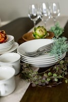 Una mesa de comedor ordenada con platos blancos, tenedores y un pequeño recipiente con comida, junto con una planta decorativa.