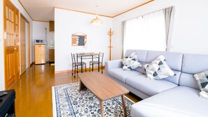 A living room with a sofa, a coffee table, a dining table with chairs, a coat rack, and a kitchen area with a refrigerator and microwave.