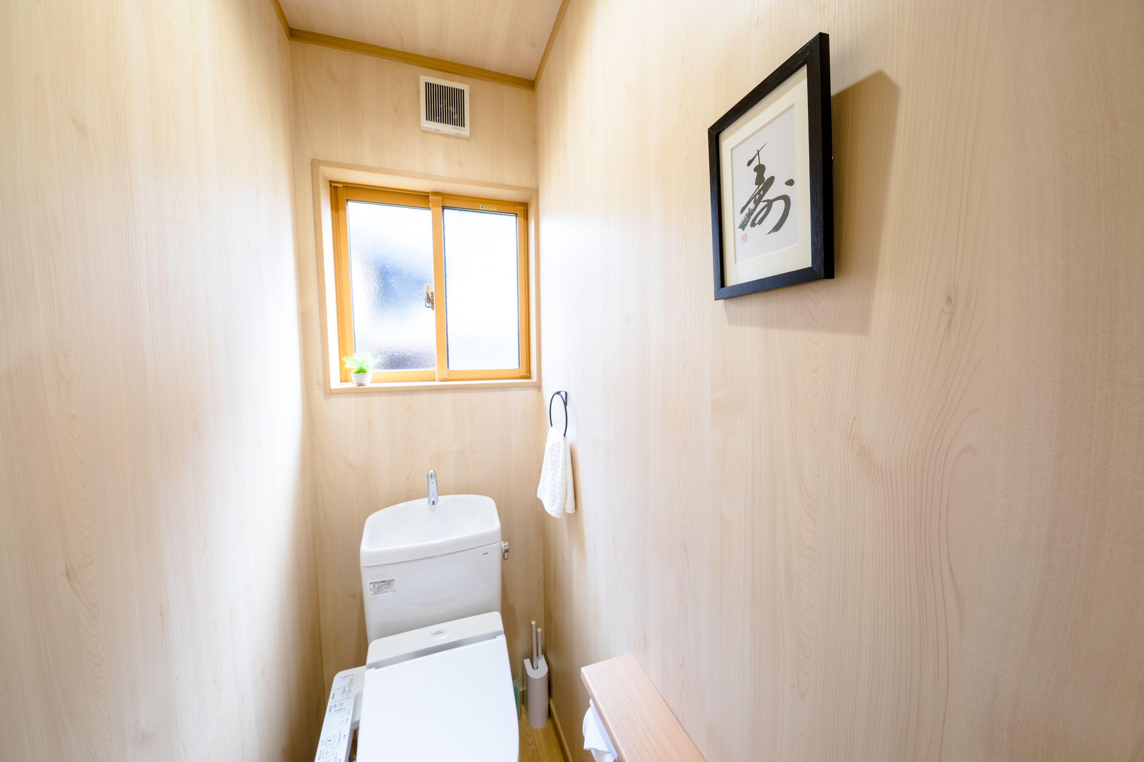 A small, wooden-finished bathroom with a toilet, a window, and a framed picture on the wall.