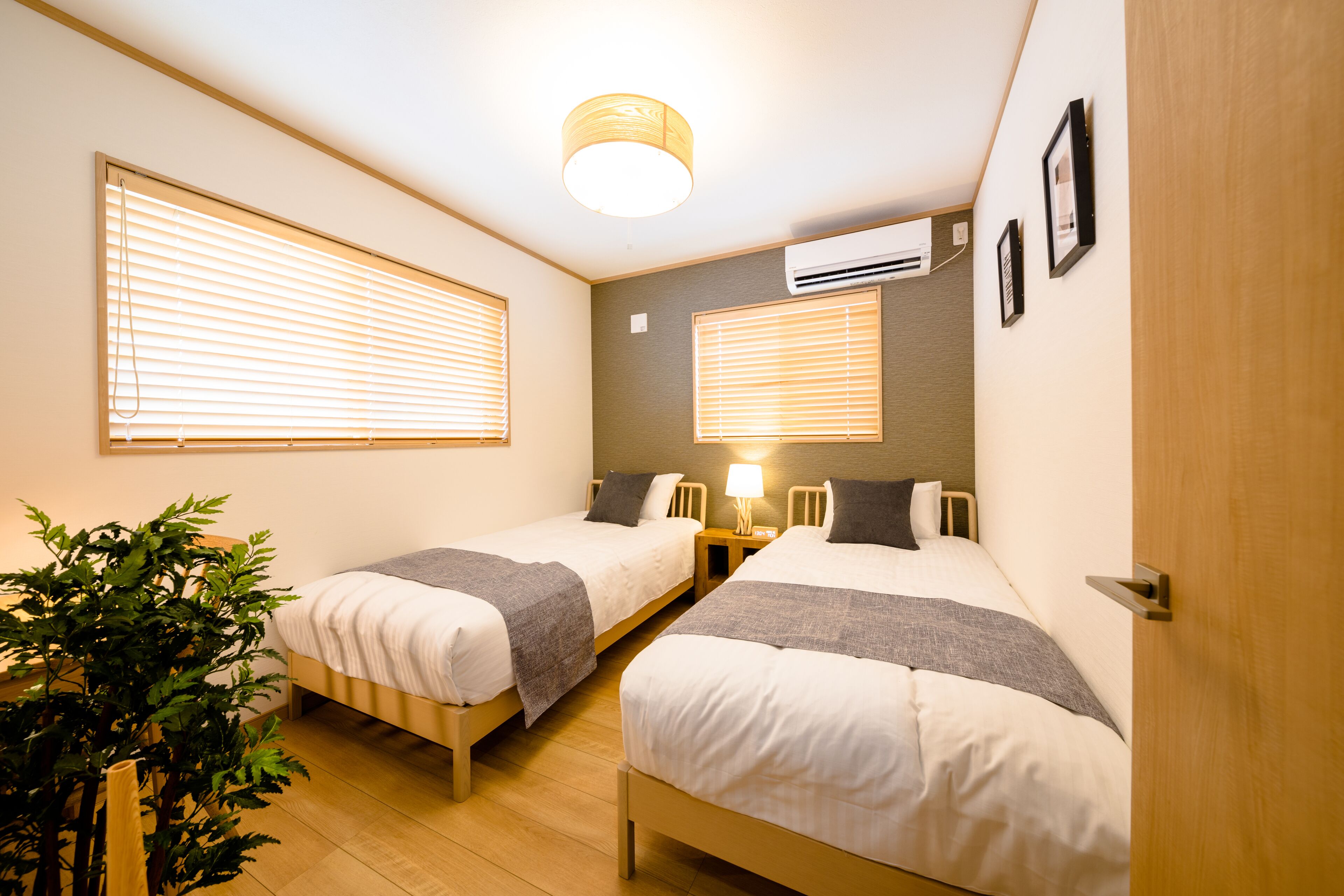 Une chambre d’hôtel avec deux lits simples, une table de chevet avec une lampe, une plante en pot et un appareil de climatisation fixé au mur.