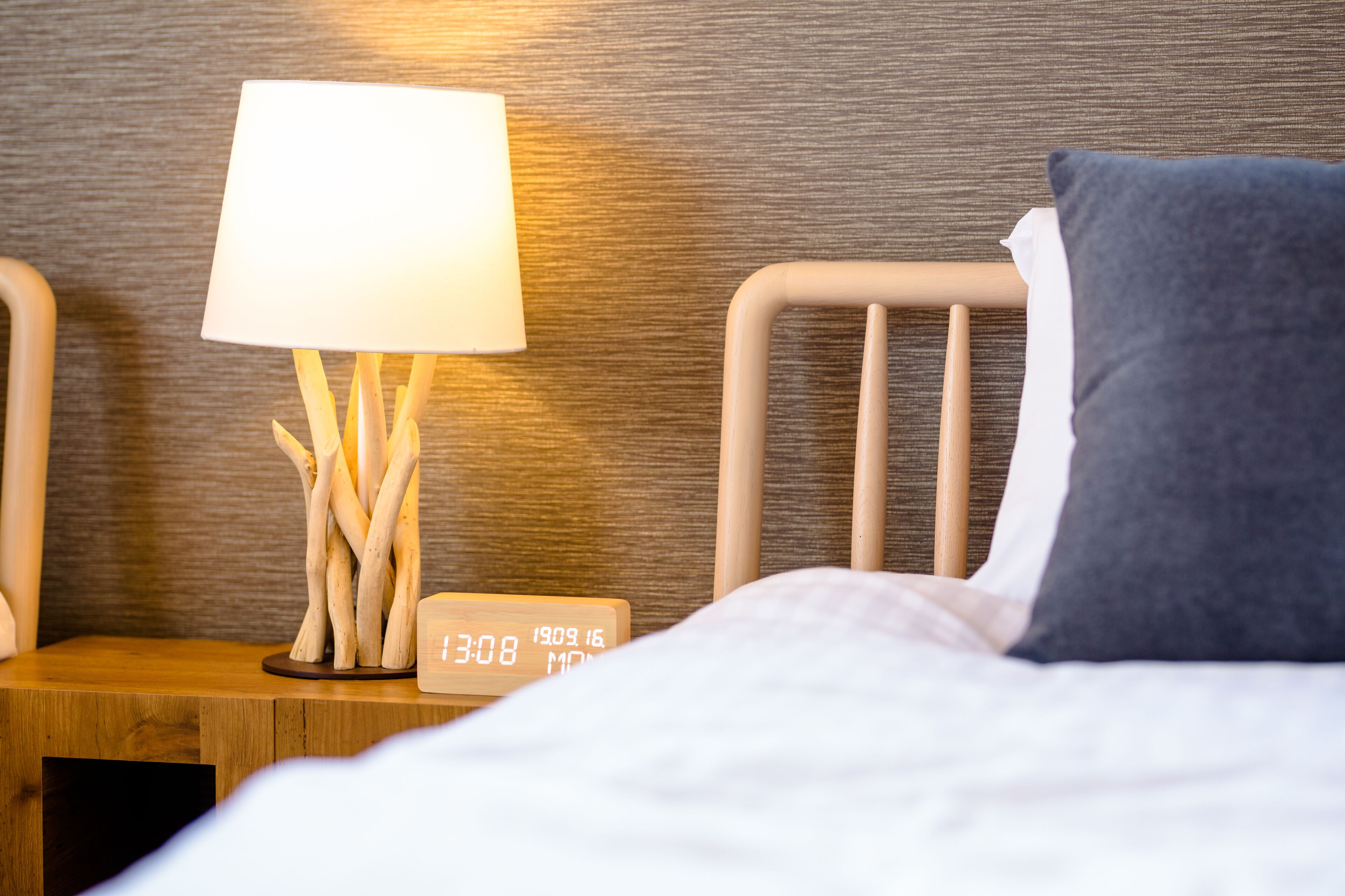Una cama bien hecha con un almohadón blanco y uno oscuro, una mesita de noche con una lámpara y un reloj, y un cabecero de madera.