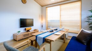 A modern living room with a wooden ceiling, a flat-screen TV, a dining table set with plates and glasses, a sofa, and a large window with blinds. - Mt. Fuji Akatsuki Enn (Fujikawaguchiko)