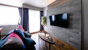 A modern living room with a flat-screen TV mounted on a brick wall, a glass coffee table, and a dark-colored sofa with cushions. - Oriya Mt.Fuji -Enishi- (Fujikawaguchiko)