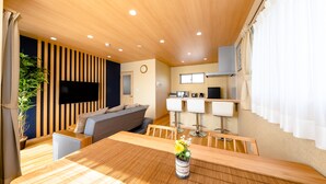 A modern living room with a wooden ceiling, a flat-screen TV mounted on the wall, a sofa, and a dining table with a vase of flowers.