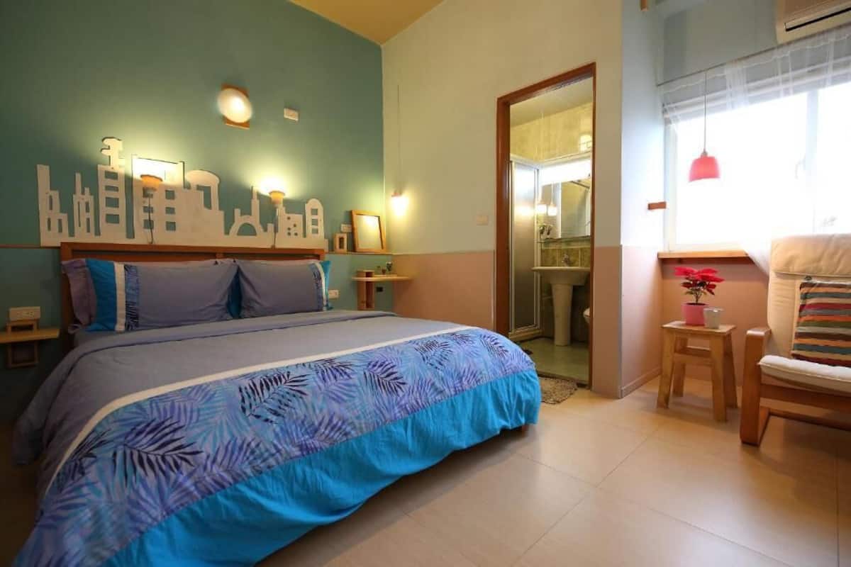A bedroom with a bed, a cityscape wall art, a small table with a plant, and a chair.