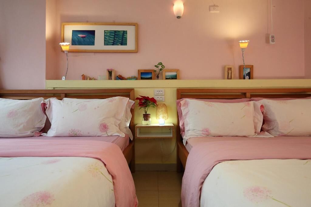 A hotel room with two beds, a nightstand with a lamp, a small table with a plant, and framed pictures on the wall.