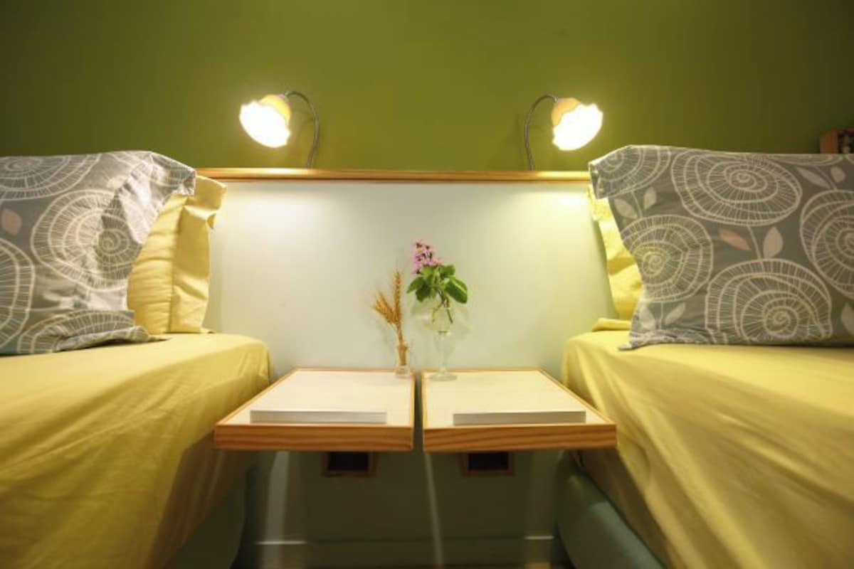 A room with two beds, each with a pillow and a bedside table. There is a small vase with flowers on the nightstand between the beds.