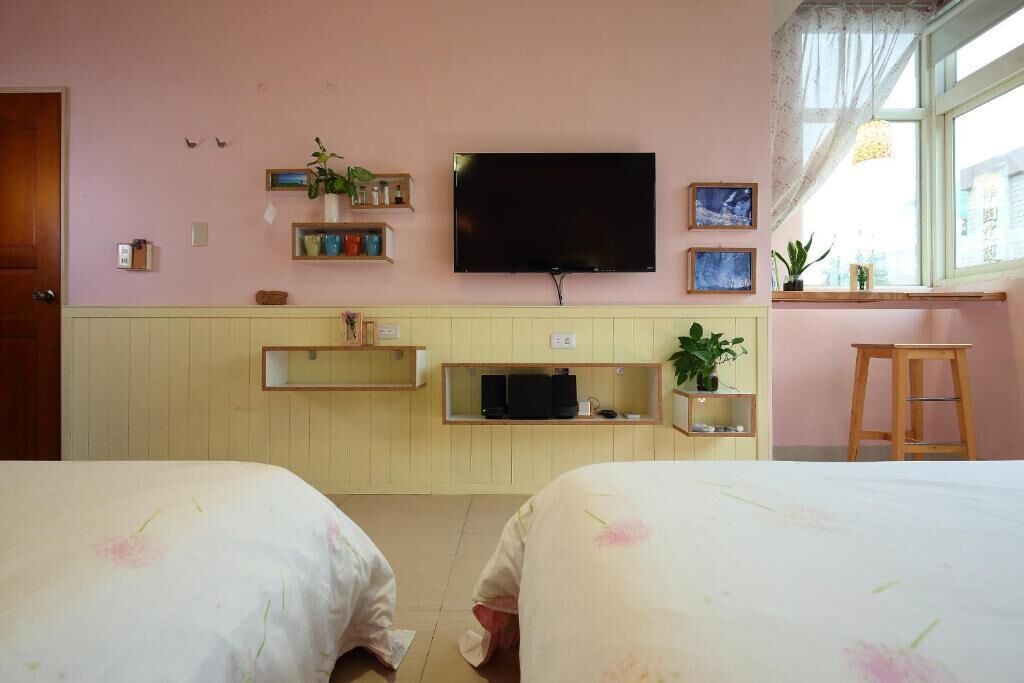 A room with two beds, a TV, shelves with plants, and a window with a view.