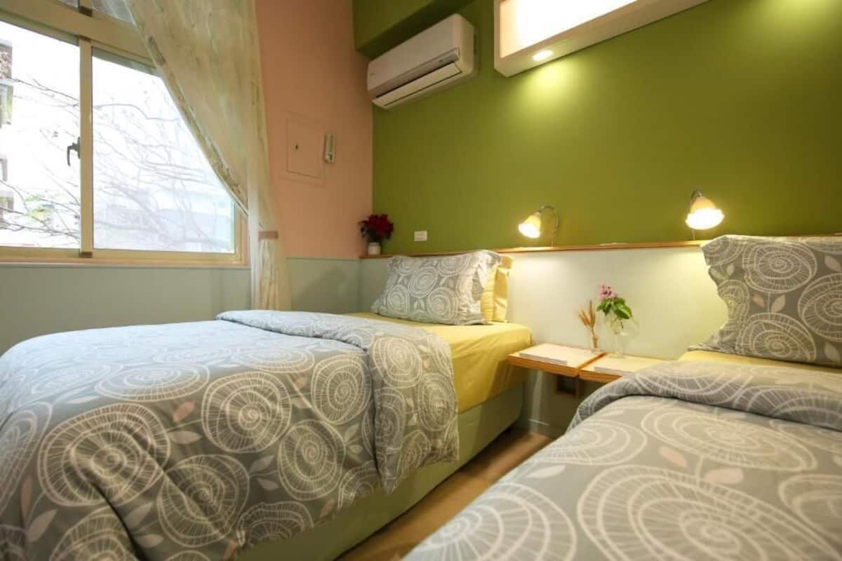 A bedroom with two beds, a window, an air conditioner, and a bedside table with a lamp.