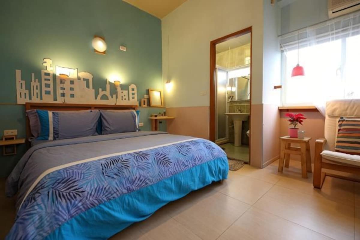 A bedroom with a bed, bedside tables, a chair, a small table with a plant, and a cityscape wall decoration.