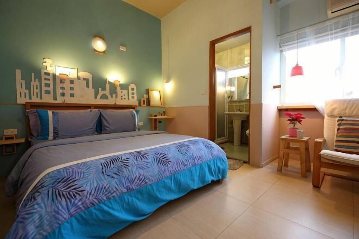 A bedroom with a bed, bedside tables, a chair, a small table with a plant, and a cityscape wall decoration.