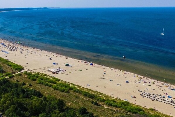 Une vue aérienne d’une plage, avec un ciel dégagé, une rive sablonneuse et un voilier à l’horizon.