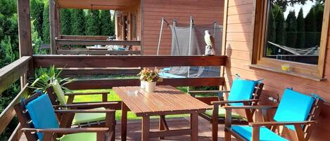 Una terraza de madera con mesa y sillas, un pequeño jardín y vista a una casa.