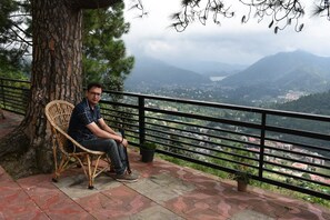 Un hombre sentado en una silla de mimbre junto a una barandilla con vistas a un valle.