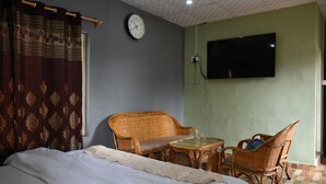 A room with a bed, two wicker chairs, a small table, a clock, and a TV on the wall. - The Moksha Heights (Nainital)