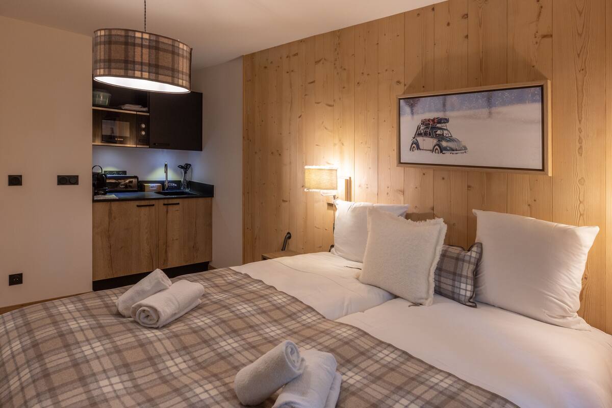 Quarto de hotel com parede de painel de madeira, cama com roupa de cama xadrez, pintura de um carro em paisagem nevada e cozinha compacta com micro-ondas e torradeira.