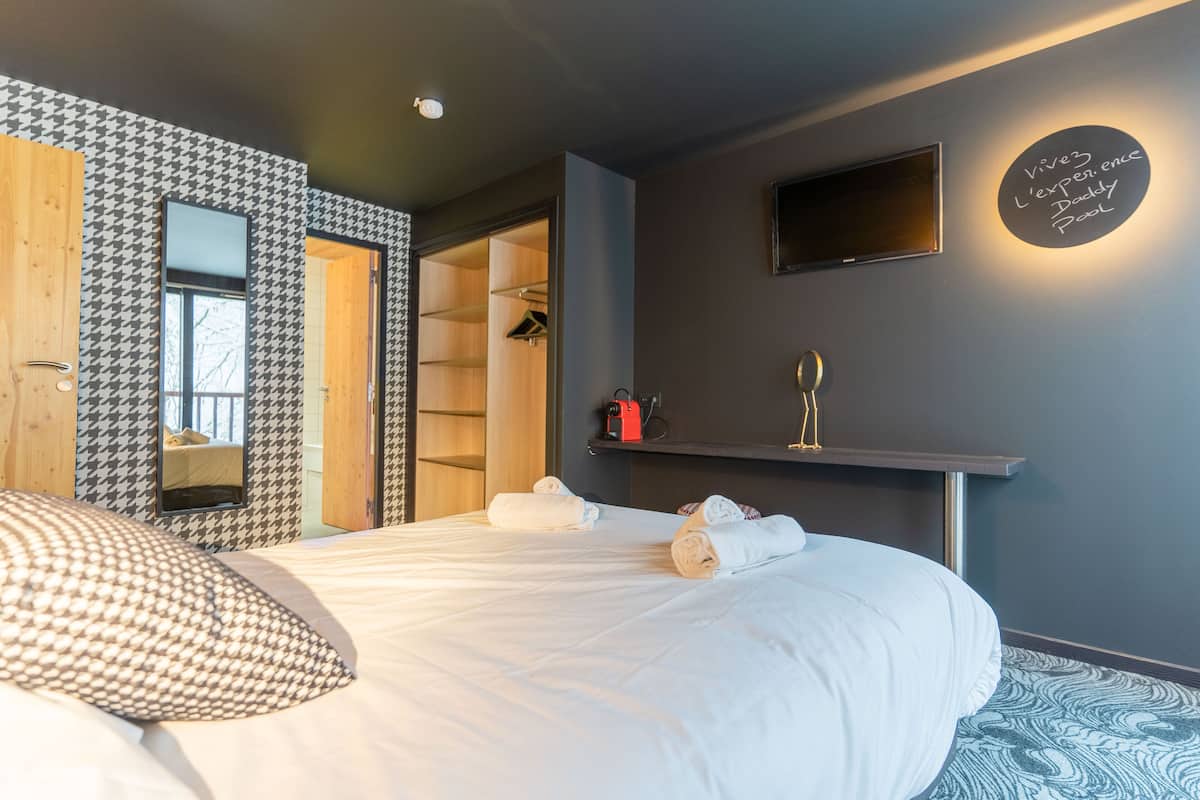 Quarto de hotel moderno com uma cama grande, um guarda-roupa, um espelho, uma TV e uma prateleira com produtos de higiene pessoal.