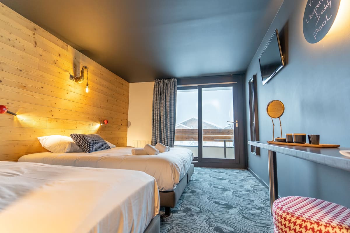 Quarto de hotel com duas camas, cabeceira de madeira, uma janela grande com vista para montanhas e uma mesinha com chávenas e um tabuleiro.
