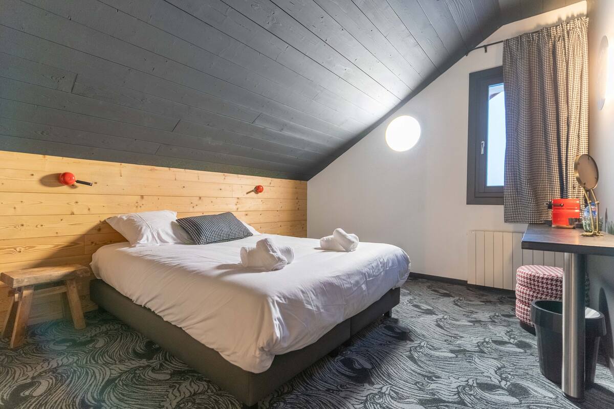 Um quarto com teto de madeira, cama com roupa de cama branca, um banquinho pequeno, uma janela com cortina e uma mesa de cabeceira com abajur e espelho.