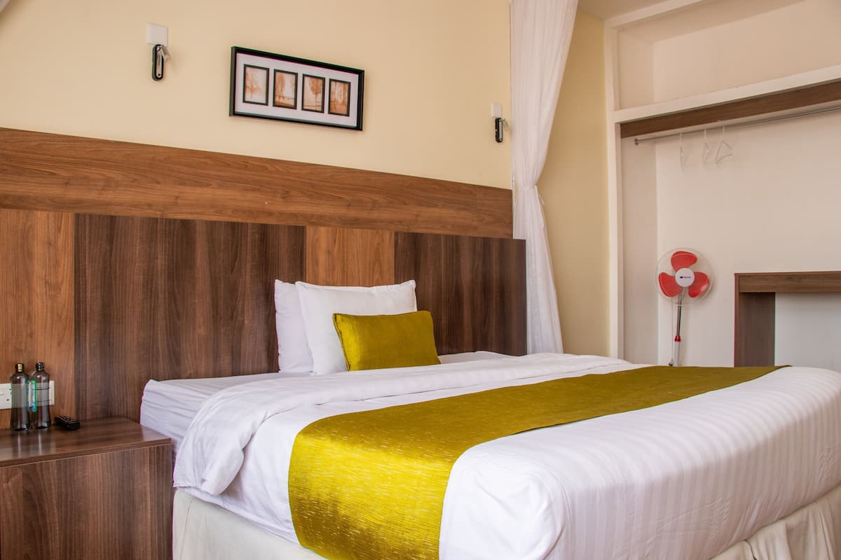A hotel room with a wooden headboard, a bed with white bedding and a gold throw, a nightstand with bottles, and a wall-mounted picture.