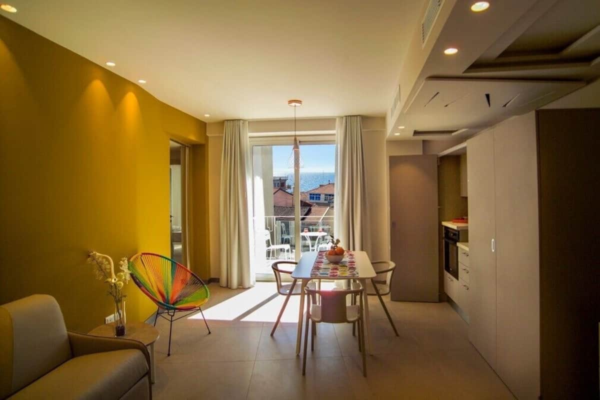 A modern living room with a dining area, a sofa, a colorful chair, and a view of the sea through large windows.