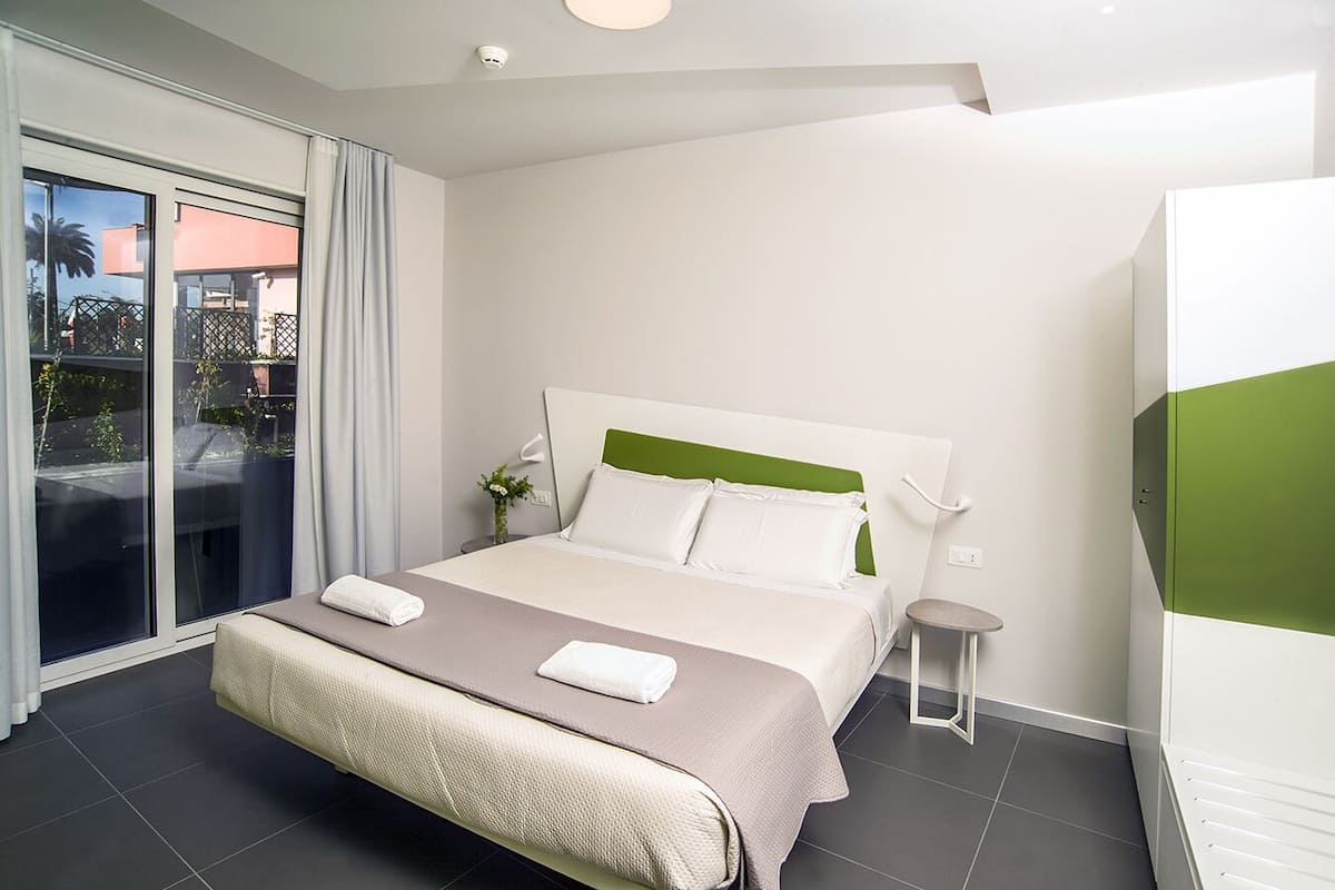 A modern hotel room with a large bed, a small bedside table, a chair, and a view of the outside.