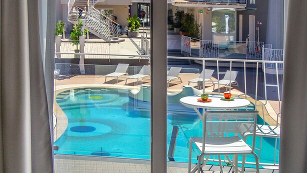 Habitación de hotel con vista a una piscina, un balcón con sillas y una mesa con una botella y fruta.