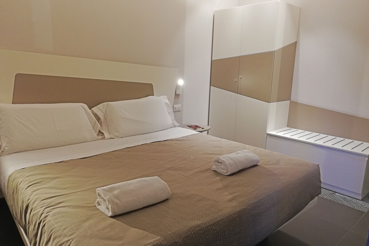 Una cama bien hecha con una colcha marrón y dos almohadas, una toalla blanca sobre la cama, y un armario con un espejo y una repisa.