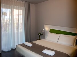 A hotel room with a large bed, a nightstand, a vase with flowers, and sheer curtains.