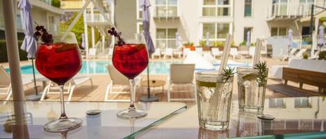 Mesa junto a la piscina con dos bebidas rojas, un vaso transparente y una botella de agua, con vista a una piscina y un edificio moderno con balcones.