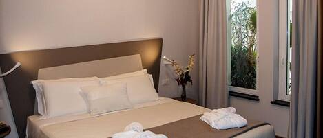 A neatly made bed with white linens and two towels, a bedside table with a vase of flowers, and a window with curtains.