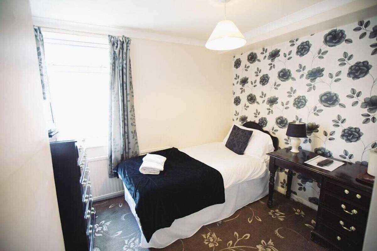 A hotel room with a bed, a bedside table with a lamp, a dresser, and a window with curtains.