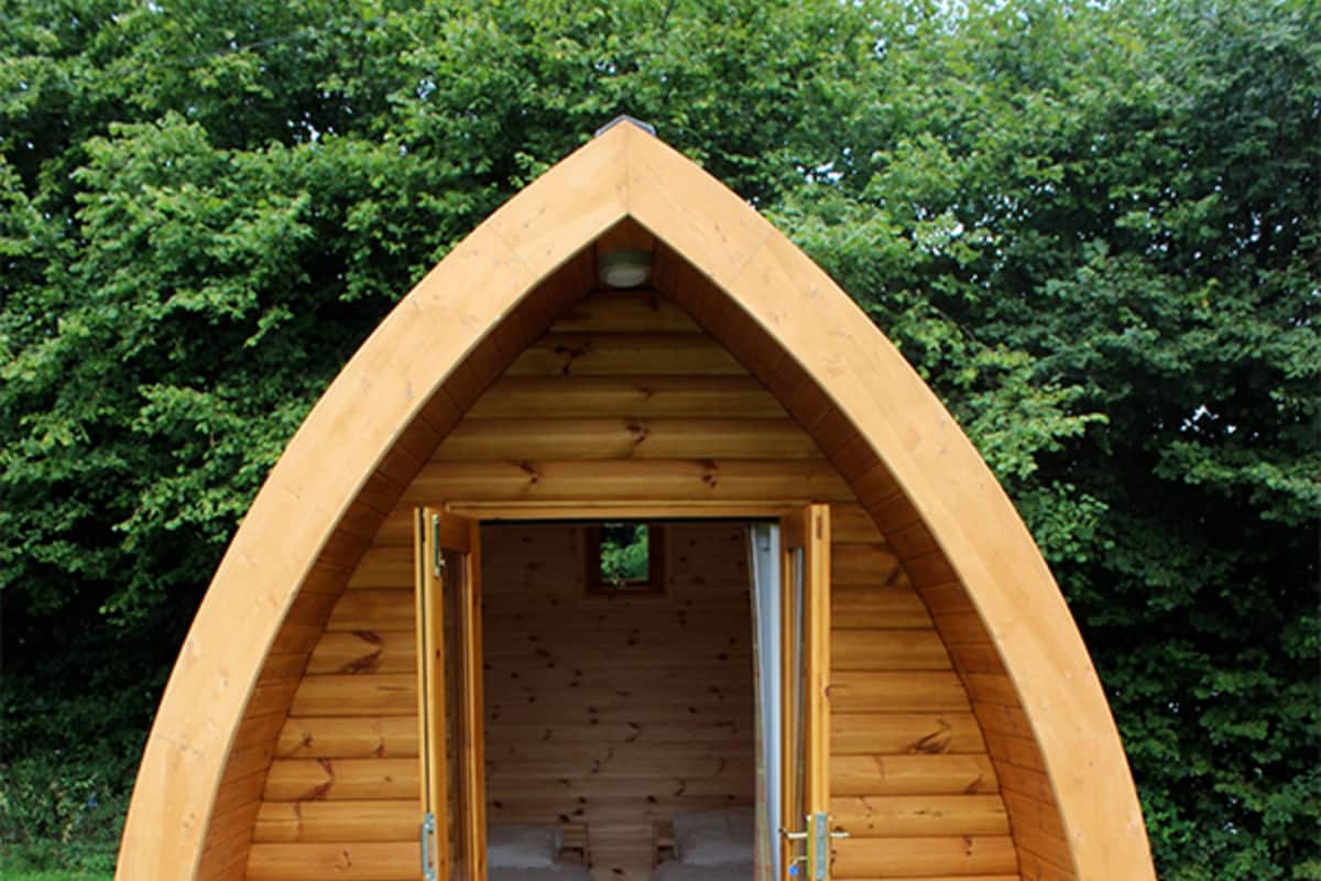 Een houten, gebogen, eenkamerhut met twee bedden en twee stoelen, omgeven door gras en bomen.