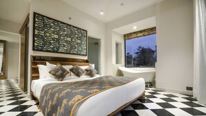Un dormitorio con piso a cuadros, una cama grande con ropa de cama estampada, un cabecero con panel decorativo, una ventana con vista a árboles, y un baño con bañera y lavamanos.