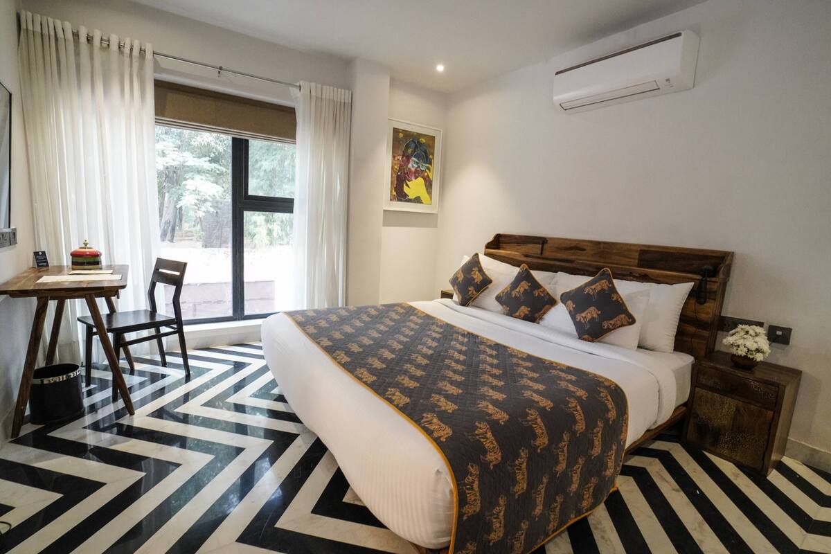 A hotel room with a bed, a desk, a chair, a window with curtains, an air conditioner, and a painting on the wall.