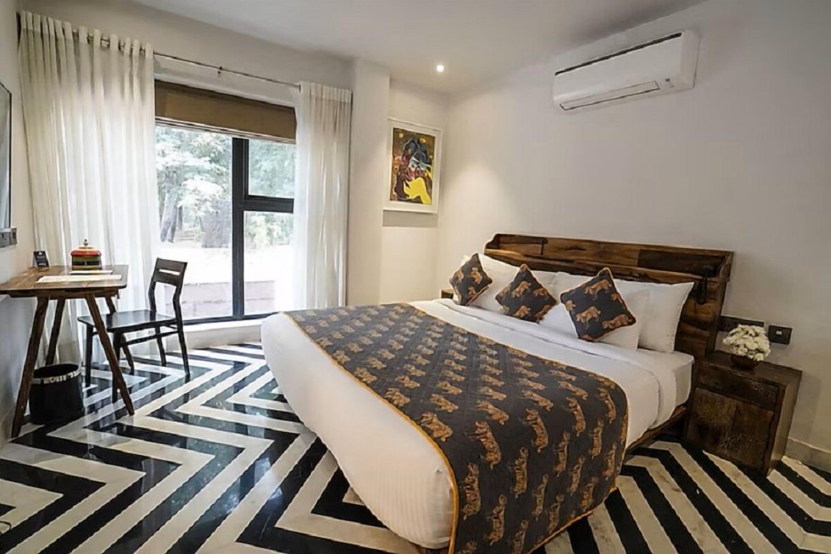 A hotel room with a large bed, a desk, and a chair. There is a window with curtains, a painting on the wall, and an air conditioning unit.