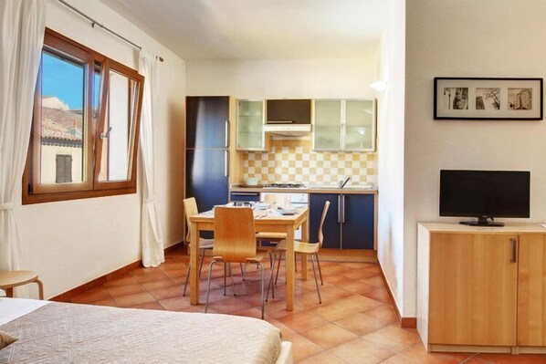 Una habitación compacta con cama, una mesa de comedor con sillas, una kitchenette con armarios y un televisor sobre un mueble.
