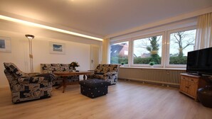 Un salon avec deux canapés à motifs, une table basse en bois, un pouf foncé, une lampe sur pied, un radiateur, un meuble pour la télévision et de grandes fenêtres donnant sur des maisons et des arbres.