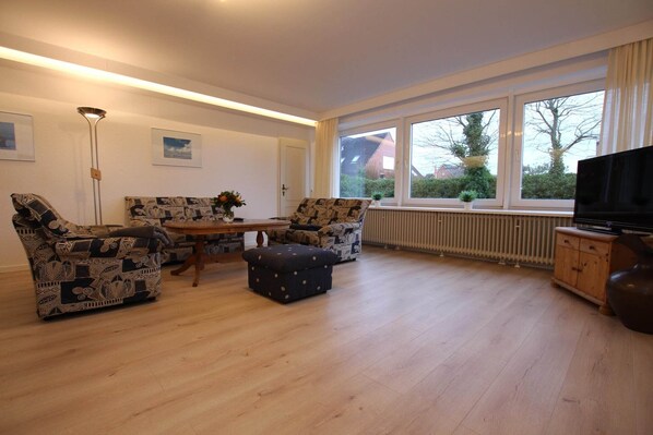 A living room with two patterned sofas, a wooden coffee table, a dark ottoman, a floor lamp, a radiator, a TV stand, and large windows with a view of houses and trees. - Ferienwohnung in Westerland auf Sylt (Westerland)