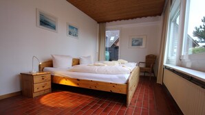 A bedroom with a wooden bed, a nightstand, a chair, and a window with a view of the outside. - Ferienwohnung in Westerland auf Sylt (Westerland)