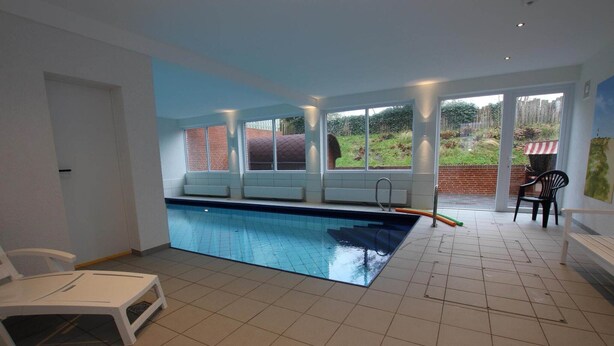 A swimming pool with a view of a brick wall and a grassy area outside. There are chairs and a table near the pool. - Ferienwohnung in Westerland auf Sylt (Westerland)