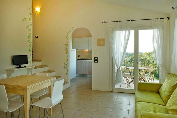 A dining area with a table and chairs, a kitchen in the background, and a balcony with a view.