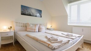 A bedroom with a bed, bedside tables, a nightstand, a window with blinds, a radiator, and a painting on the wall.