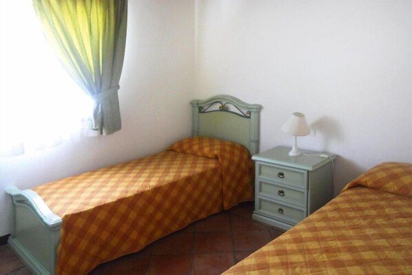 Une petite chambre avec un lit simple, une couverture à carreaux, une table de chevet avec une lampe et une fenêtre avec des rideaux.